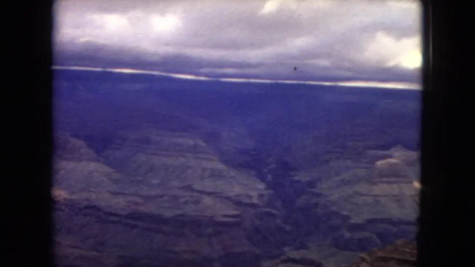 Final 1967 8mm frame showing a broad Grand Canyon vista, with deep blue canyon shadows, a pale horizon line, cloud cover, and rocky foreground at the lower edge.