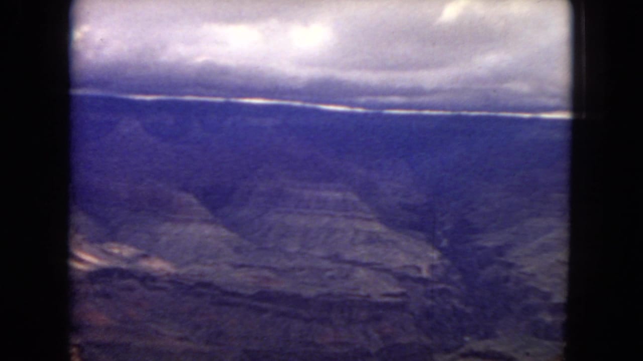 1967 8mm Arizona home-movie frame showing layered Grand Canyon plateaus under a dense cloud bank, with soft color shift and dark gate edges.