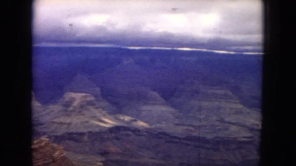 1967 8mm Arizona frame looking across Grand Canyon formations and distant plateaus beneath rolling clouds and muted blue-purple film color.