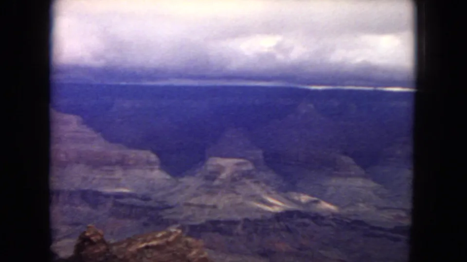 1967 8mm home-movie frame from an Arizona canyon overlook, showing mesas, ridges, and a low overcast sky with visible film grain and edge shadow.