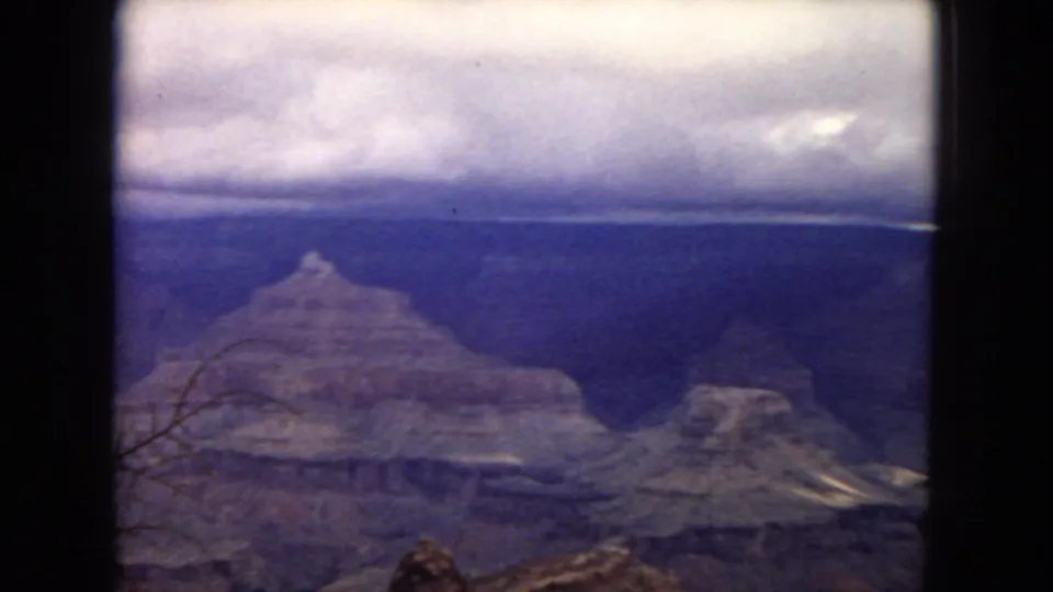 1967 8mm Arizona frame showing Grand Canyon plateaus under a dark cloud layer, with a small foreground rock and bare branch visible near the lower edge.