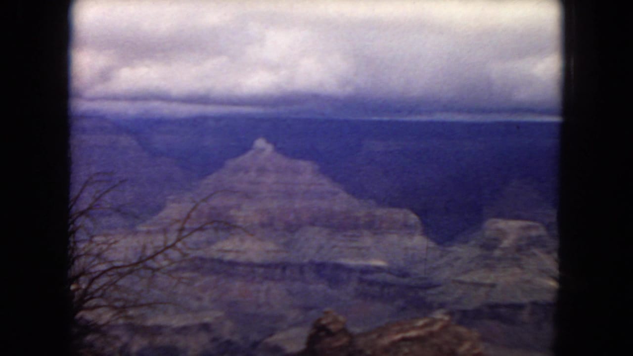 1967 8mm home-movie frame of a broad Arizona canyon view with layered rock formations, a cloudy sky, and sparse foreground brush.