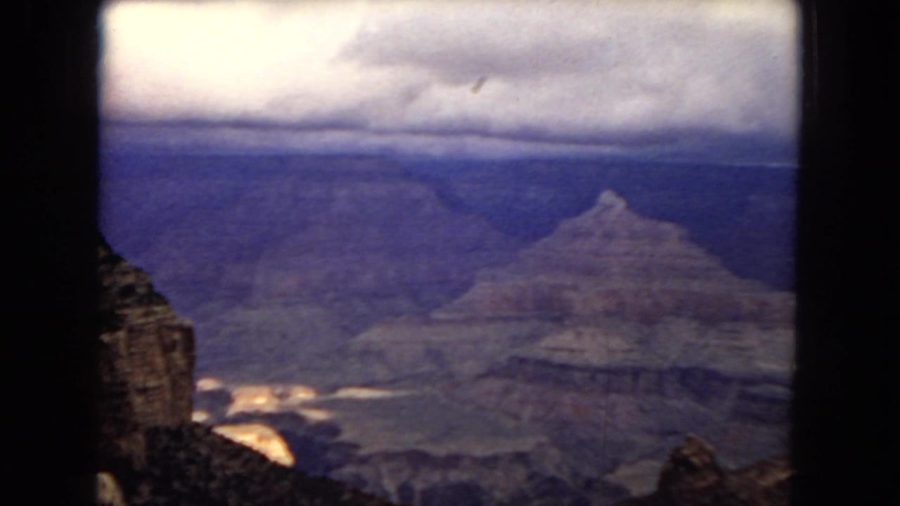 1967 8mm Arizona canyon view with a dark rock face in the foreground, distant stratified walls, and bright light catching part of the gorge.