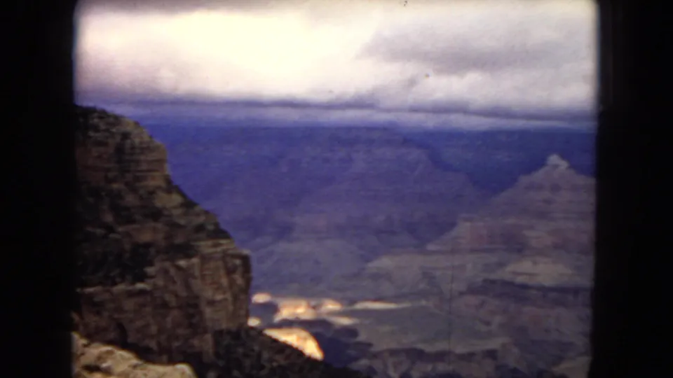 1967 8mm home-movie frame looking past a dark canyon rim toward blue-toned cliffs and a cloudy horizon in Arizona.
