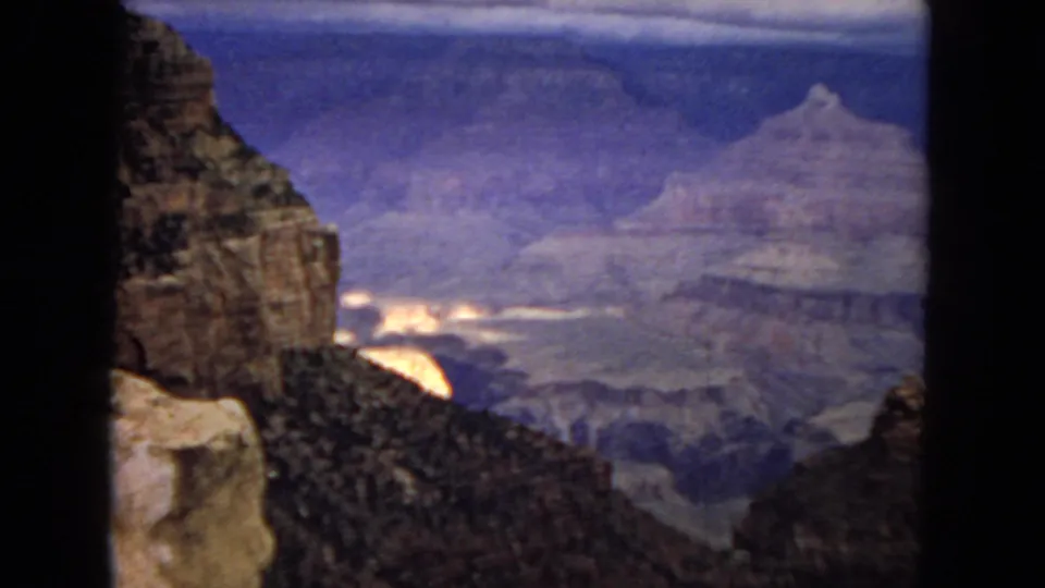 1967 8mm Arizona canyon frame showing a dark foreground cliff, a deep gorge, distant layered rock, and overcast light across the landscape.