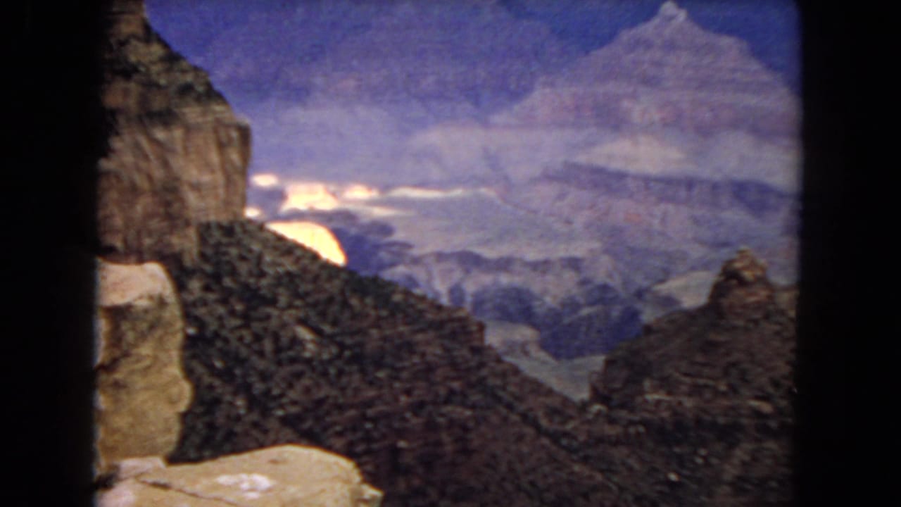 1967 8mm home-movie frame of the Grand Canyon with dark foreground ridges, pale rock ledges, distant layered canyon walls, and blue sky.