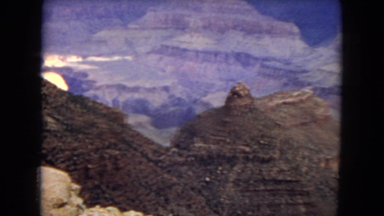 1967 8mm home-movie frame looking across the Grand Canyon, with a dark rocky foreground ridge, distant plateau layers, and a small sunlit rock area near the horizon.