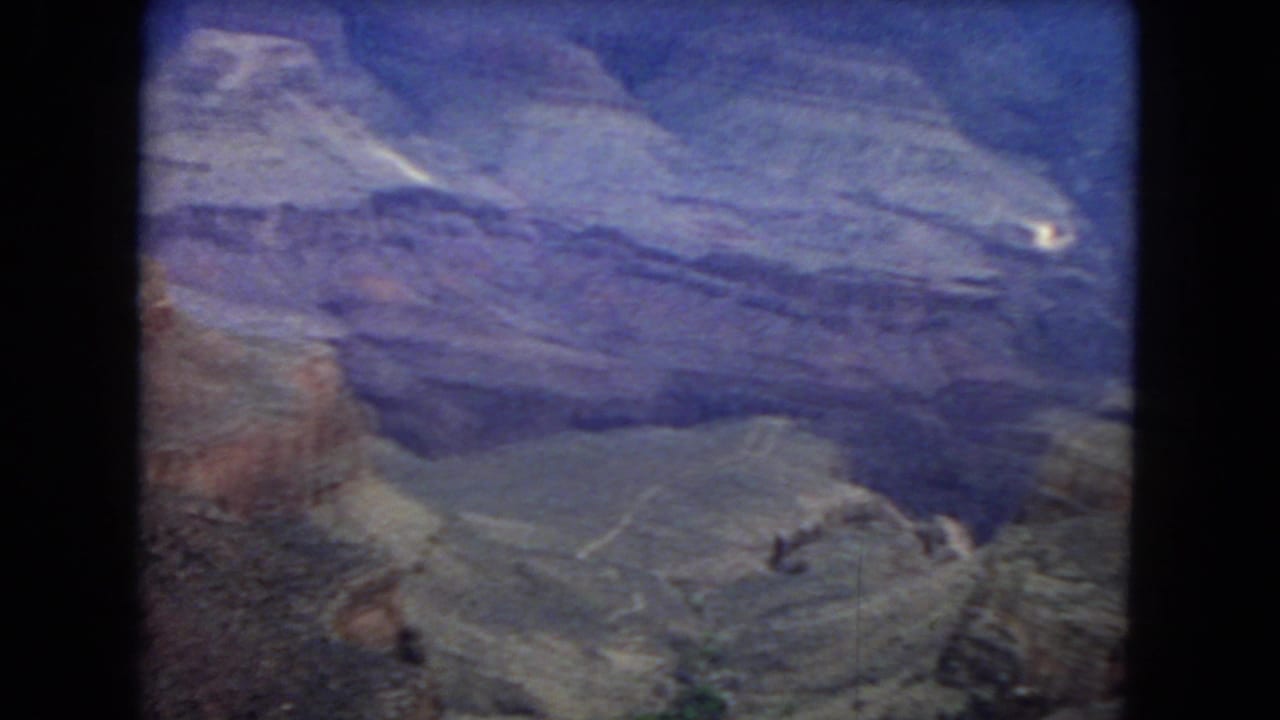 1967 8mm home-movie frame showing the Grand Canyon from above, with layered rock walls, deep ravines, and a purple-blue aged film cast.