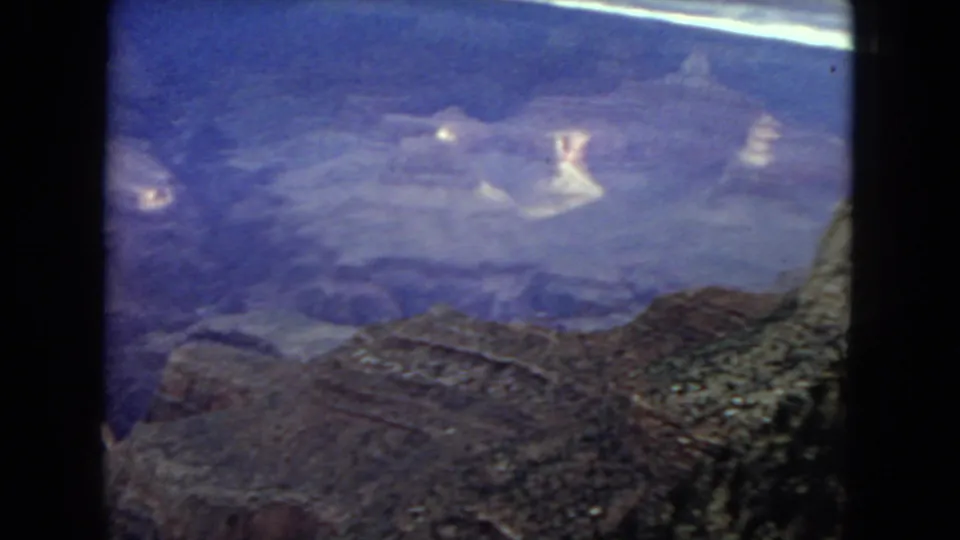 Aged 8mm frame of the Grand Canyon panorama, showing bluish faded film color, distant canyon walls, and darker foreground rock along the lower edge.