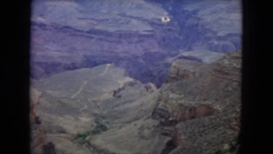1967 8mm frame looking down into the Grand Canyon, with sloped rock shelves, layered cliffs, and a prominent foreground formation on the right.
