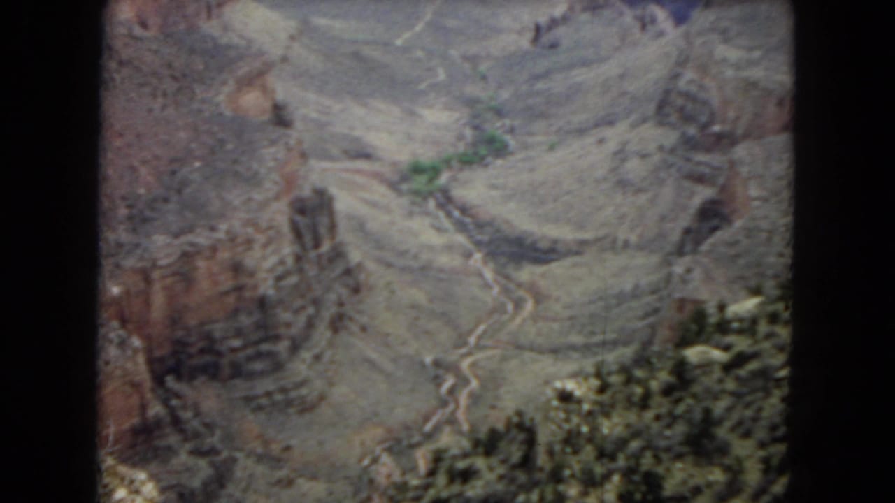 A vintage 8mm frame looks down into the Arizona canyon terrain, showing red-brown cliffs, gray slopes, a winding channel, and scattered greenery.