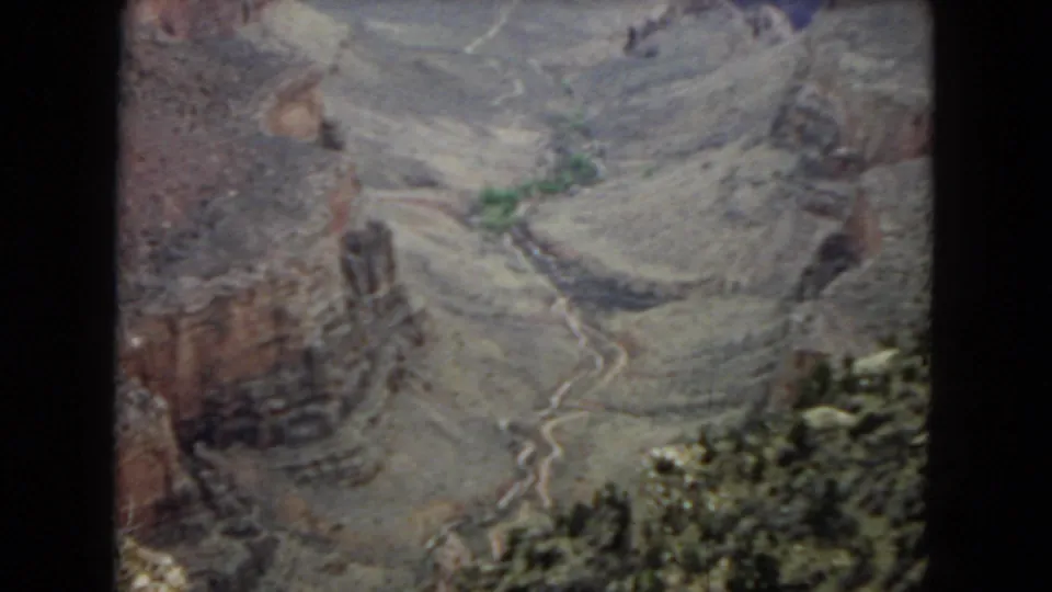 A vintage 8mm frame looks down into the Arizona canyon terrain, showing red-brown cliffs, gray slopes, a winding channel, and scattered greenery.