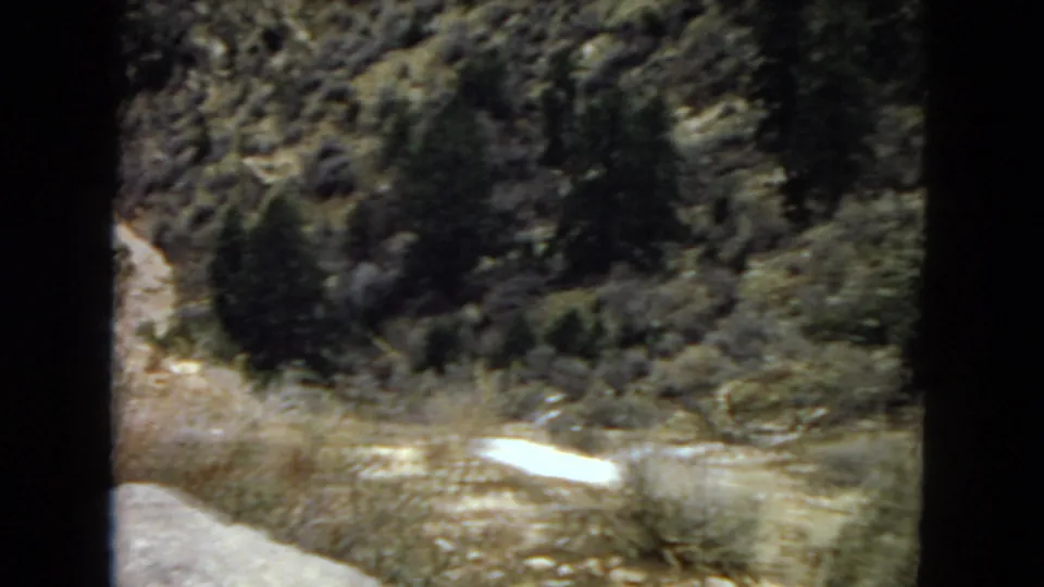 1967 Arizona desert landscape frame with dark shrubs, rocky ground, and a pale strip of dry wash or exposed stone along the lower slope.