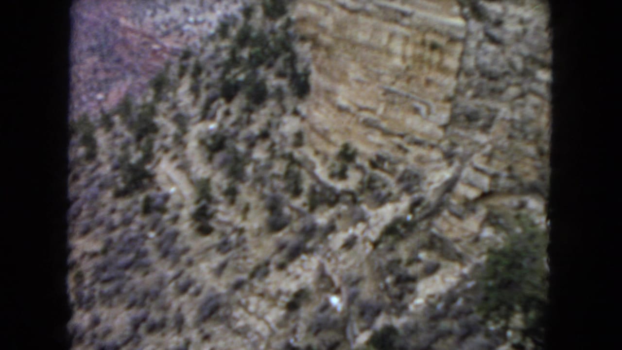 1967 Arizona 8mm frame showing a steep Grand Canyon rock wall, broken ledges, sparse vegetation, and a soft handheld view through dark film borders.