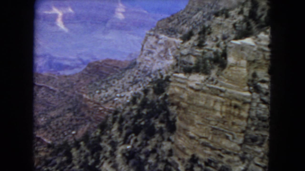 1967 Arizona 8mm frame looking across Grand Canyon cliffs toward distant ridges under a deep blue sky with scattered light cloud marks.