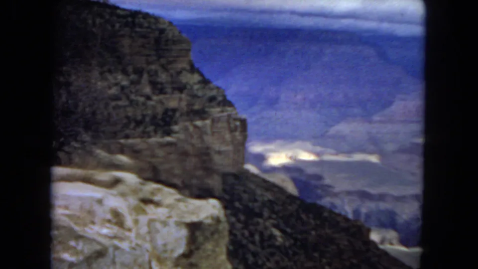 1967 Arizona 8mm frame with a foreground stone ledge, dark canyon wall, hazy blue distance, and pale sunlit areas far below.