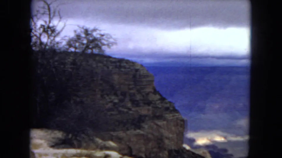 1967 Arizona 8mm frame showing a shadowed canyon rim, sparse bare branches, layered rock, and a cloudy blue sky over the distant canyon.
