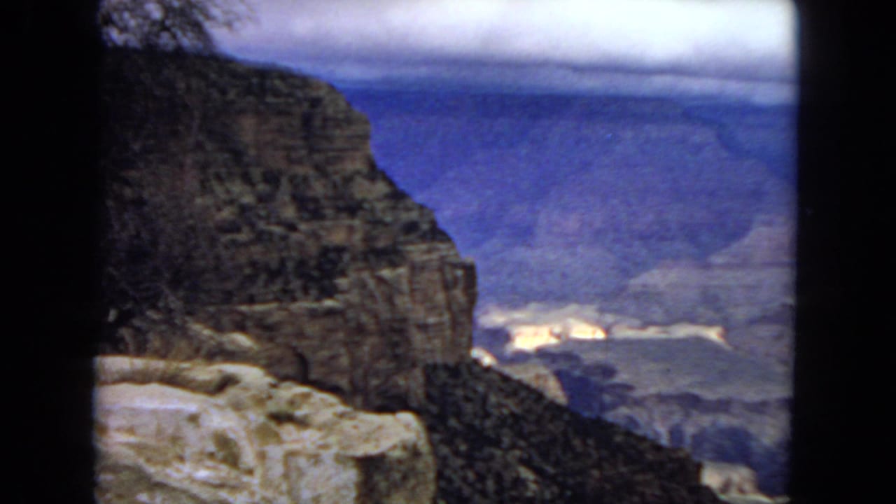 1967 8mm view of the Grand Canyon from a rocky overlook, with dark foreground cliffs and hazy layered canyon walls in the distance.