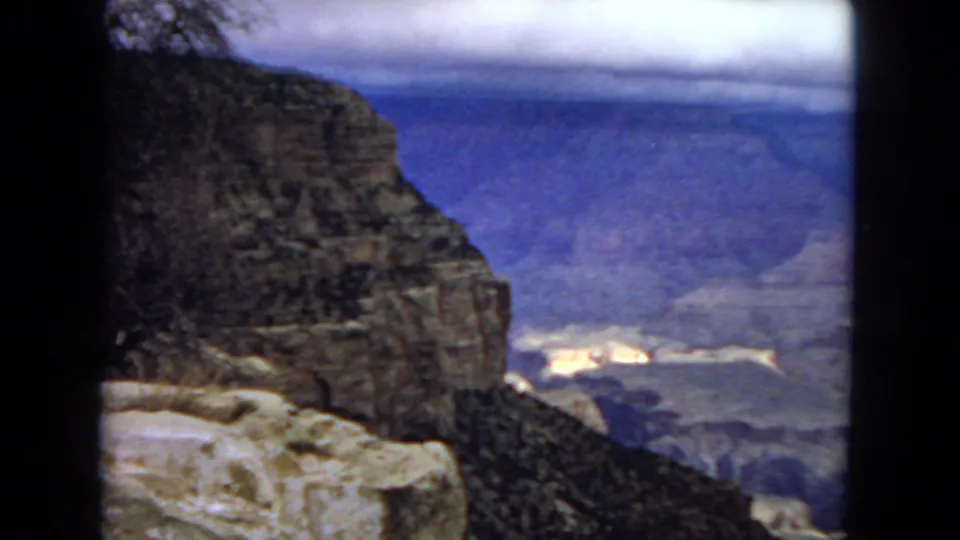 1967 8mm view of the Grand Canyon from a rocky overlook, with dark foreground cliffs and hazy layered canyon walls in the distance.