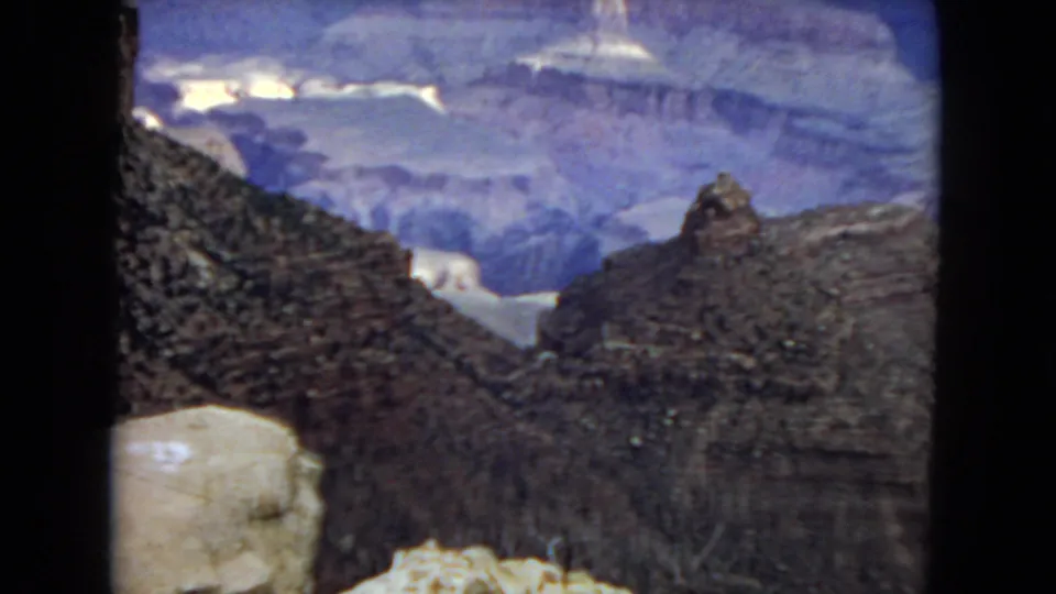 Visitor-shot 8mm frame of the Grand Canyon showing a dark rocky slope, pale stone ledges, and distant blue-toned canyon layers.