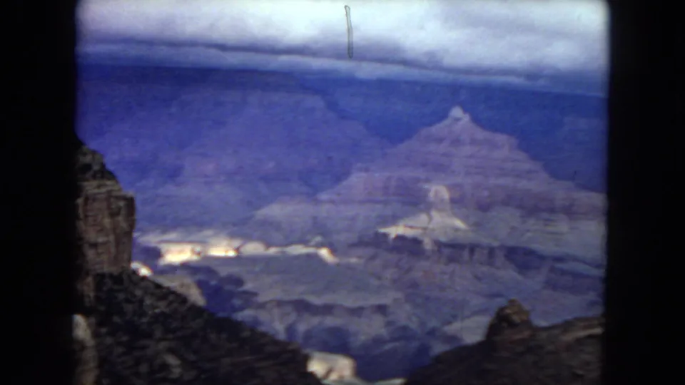 Wide 1967 Grand Canyon view with foreground rock shapes, distant mesas, cloudy sky, and strong blue-purple fading from the 8mm film scan.