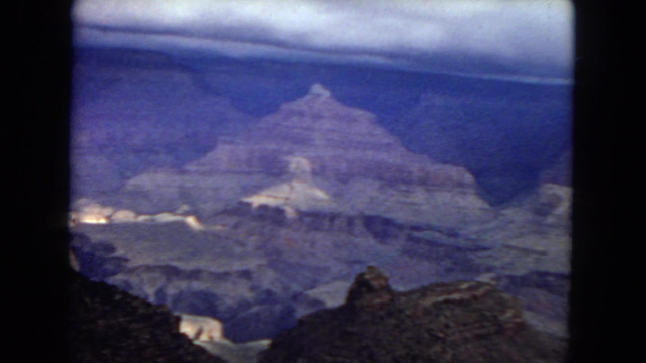 1967 8mm home-movie frame showing a Grand Canyon overlook view with layered rock walls, a dark foreground ledge, and a purple-blue aged-film cast.
