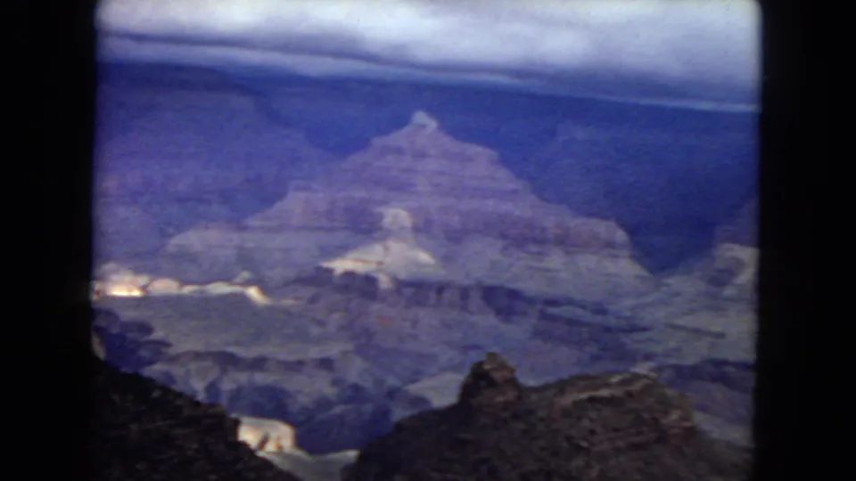 1967 8mm home-movie frame showing a Grand Canyon overlook view with layered rock walls, a dark foreground ledge, and a purple-blue aged-film cast.