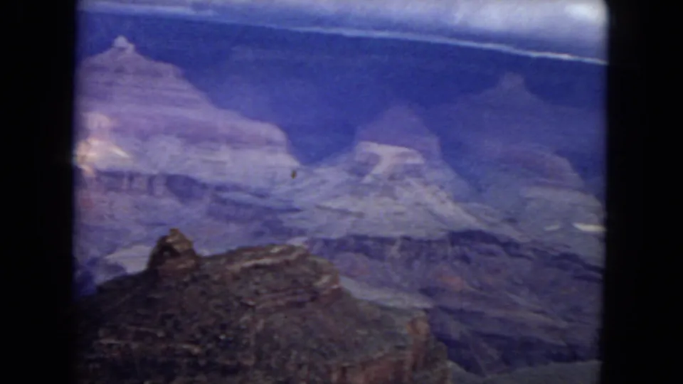 1967 8mm frame of the Grand Canyon panorama with broad blue-purple shadows, stepped rock formations, and a dark rocky foreground edge.