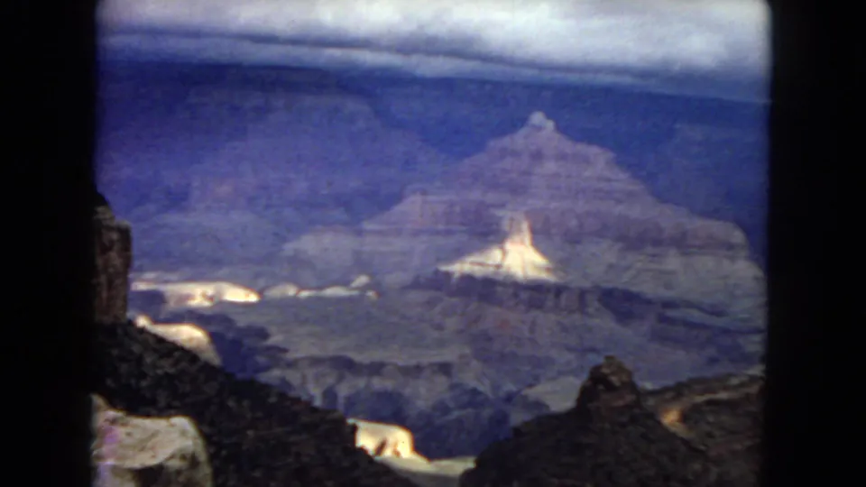 1967 8mm home-movie frame looking across the Grand Canyon from a rocky rim, with pale stone shelves, deep shadowed layers, and aged film grain.