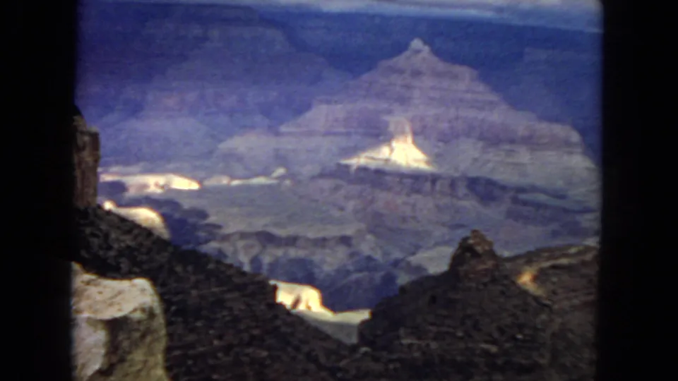 1967 8mm frame showing a wide Grand Canyon vista with foreground boulders, bright exposed rock, distant cliff bands, and heavy blue-purple film aging.