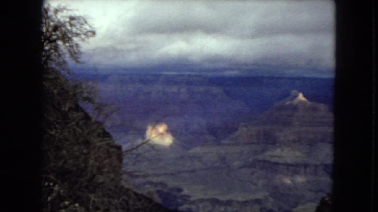 1967 8mm view of the Grand Canyon with layered blue-gray canyon walls, cloudy sky, dark foreground trees, and a small sunlit rock formation.