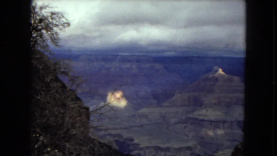 1967 8mm view of the Grand Canyon with layered blue-gray canyon walls, cloudy sky, dark foreground trees, and a small sunlit rock formation.