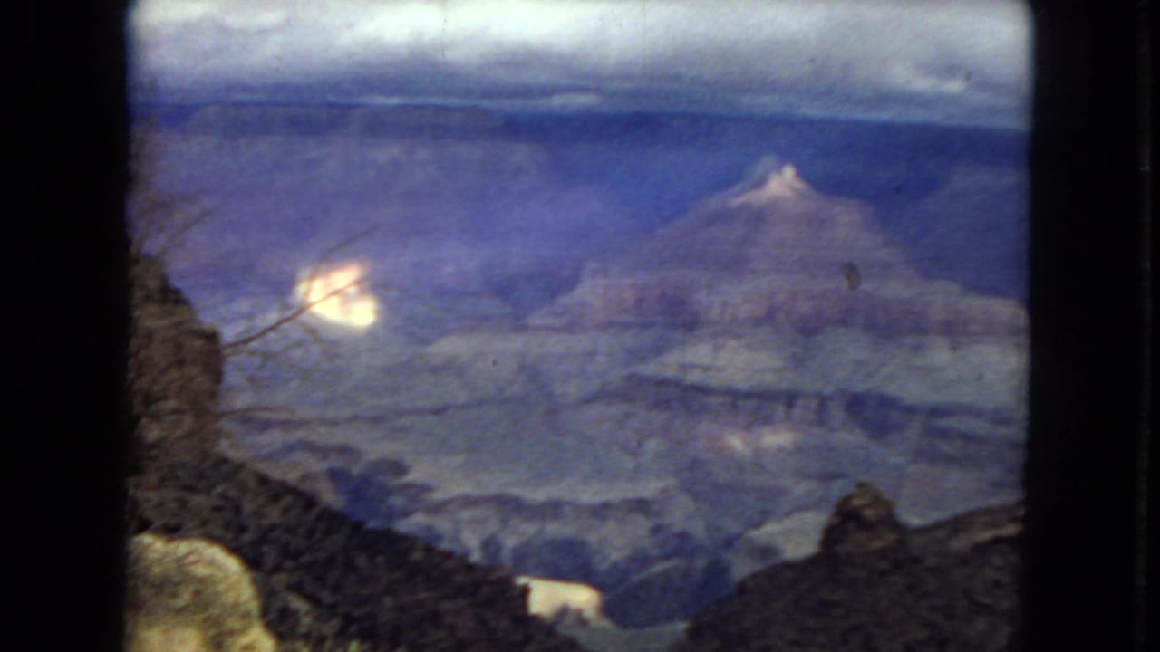 1967 8mm Grand Canyon panorama showing a cloudy skyline, layered canyon formations, a dark foreground edge, and bright patches of sunlight on rock.