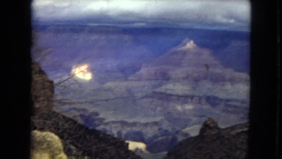 1967 8mm Grand Canyon panorama showing a cloudy skyline, layered canyon formations, a dark foreground edge, and bright patches of sunlight on rock.