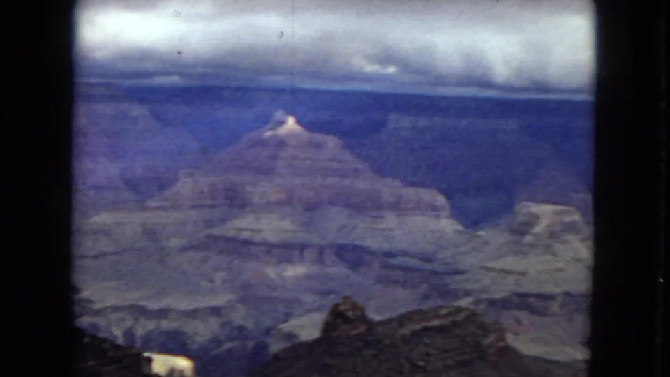1967 8mm Grand Canyon landscape with a central rock formation, layered canyon walls, cloudy sky, and dark rim rocks in the foreground.