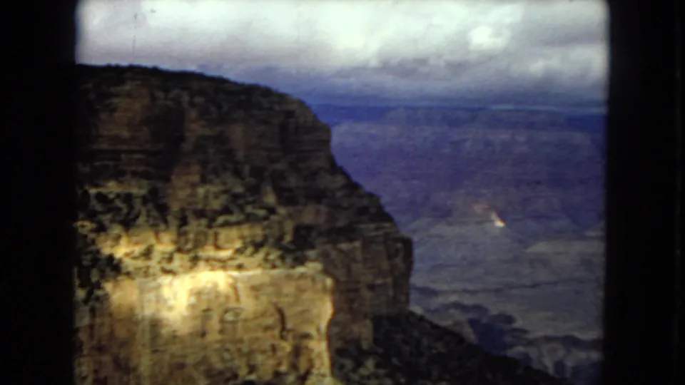 1967 8mm view from the Grand Canyon rim, with a dark foreground cliff face, distant blue-gray canyon layers, and a cloudy horizon.