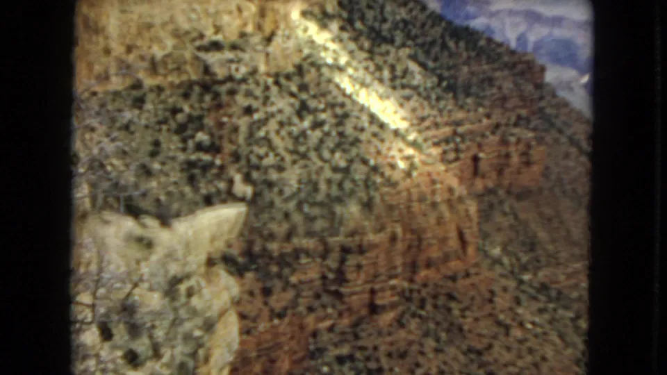1967 Arizona 8mm frame showing a closer view of a canyon slope, pale foreground rocks, reddish layered cliff bands, and a bright diagonal area of sunlight.