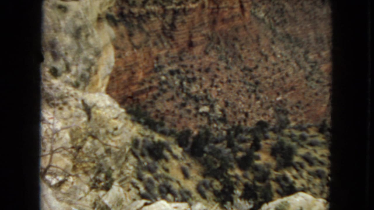 1967 Arizona 8mm frame showing a rocky canyon wall with pale foreground stone, scattered shrubs, and warm red-brown rock tones.