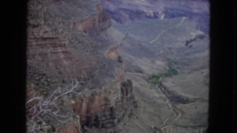 Grand Canyon panorama from a visitor overlook, showing layered cliffs, pale winding terrain, sparse branches, and atmospheric haze in 1967 8mm color.