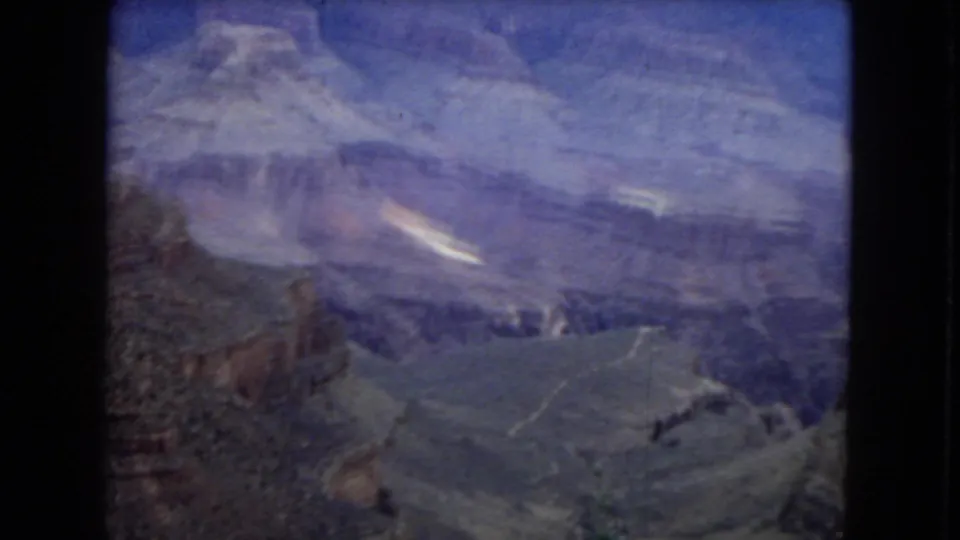 Wide 8mm Grand Canyon view with distant ridges, broad canyon depth, muted rock layers, and a soft purple-blue cast from the film stock.