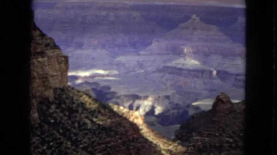 1967 Grand Canyon home-movie frame with a dark cliff edge in the foreground, sunlit rock highlights, distant canyon formations, and heavy atmospheric haze.
