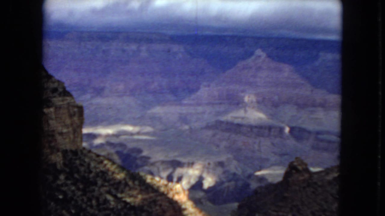 1967 8mm home-movie frame of the Grand Canyon, showing layered canyon walls, a foreground slope, and dark clouds above the distant rim.