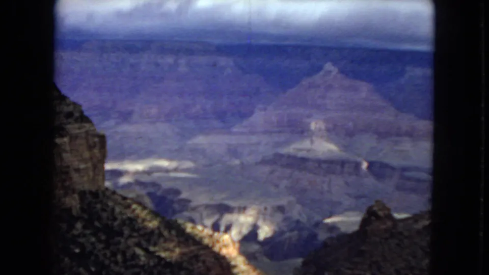 1967 8mm home-movie frame of the Grand Canyon, showing layered canyon walls, a foreground slope, and dark clouds above the distant rim.