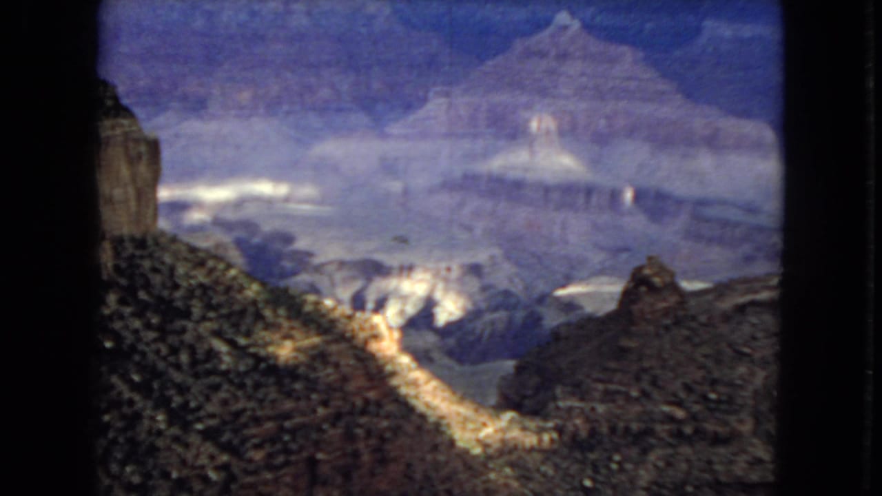 1967 Arizona home-movie view across the Grand Canyon, with rocky foreground terrain, distant mesas, and uneven light beneath a cloudy sky.