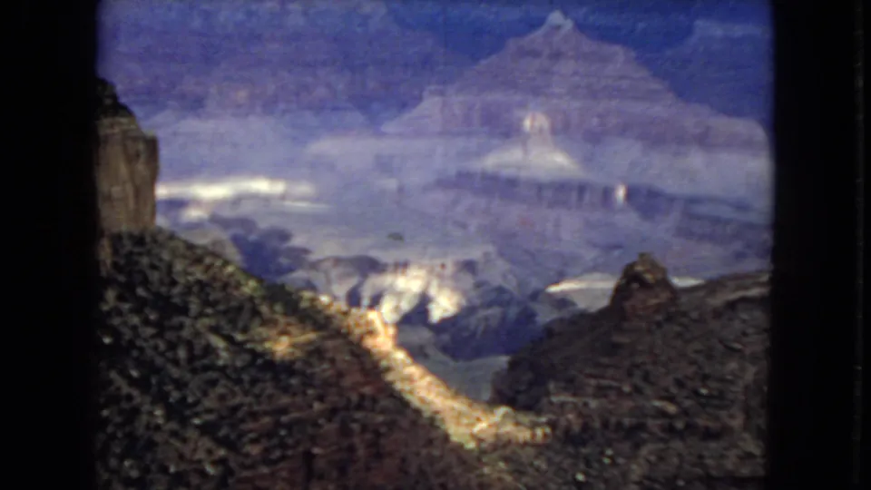 1967 Arizona home-movie view across the Grand Canyon, with rocky foreground terrain, distant mesas, and uneven light beneath a cloudy sky.