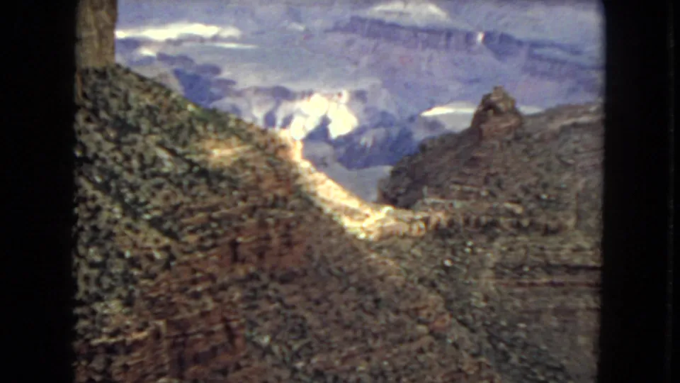 1967 8mm frame looking into the Grand Canyon, with a steep foreground slope, dark ravine forms, and a bright patch of exposed rock in the distance.