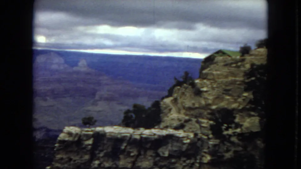 1967 Grand Canyon home-movie frame showing a rocky rim or overlook in the foreground, sparse dark vegetation, and a broad blue-gray canyon under low clouds.