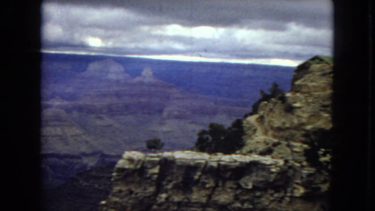 1967 Arizona 8mm frame showing distant canyon walls, a pale stratified ledge, sparse shrubs, and a moody overcast sky from a high overlook.