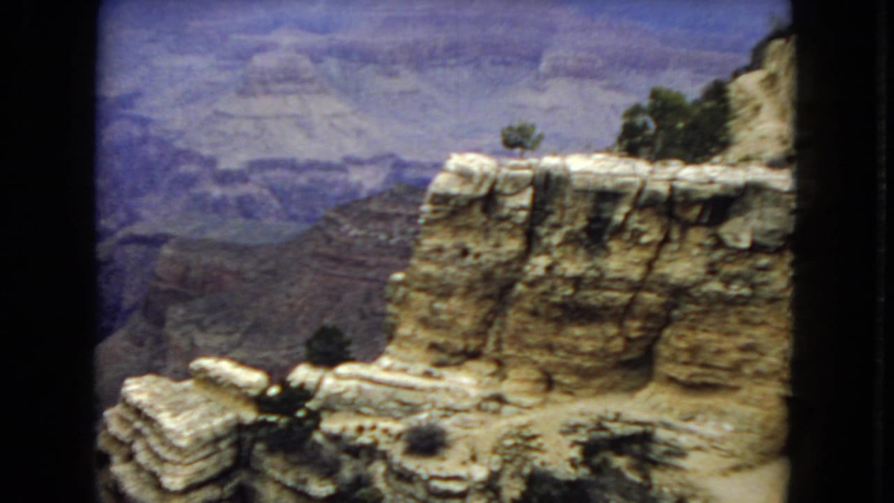 1967 Arizona 8mm frame with a pale layered rock shelf, scrub vegetation, and hazy canyon formations receding in the background.