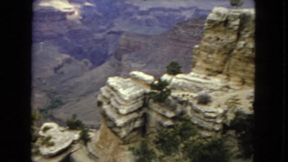 1967 Arizona 8mm frame looking across layered sandstone-like ledges and desert shrubs toward broad canyon walls in soft focus.